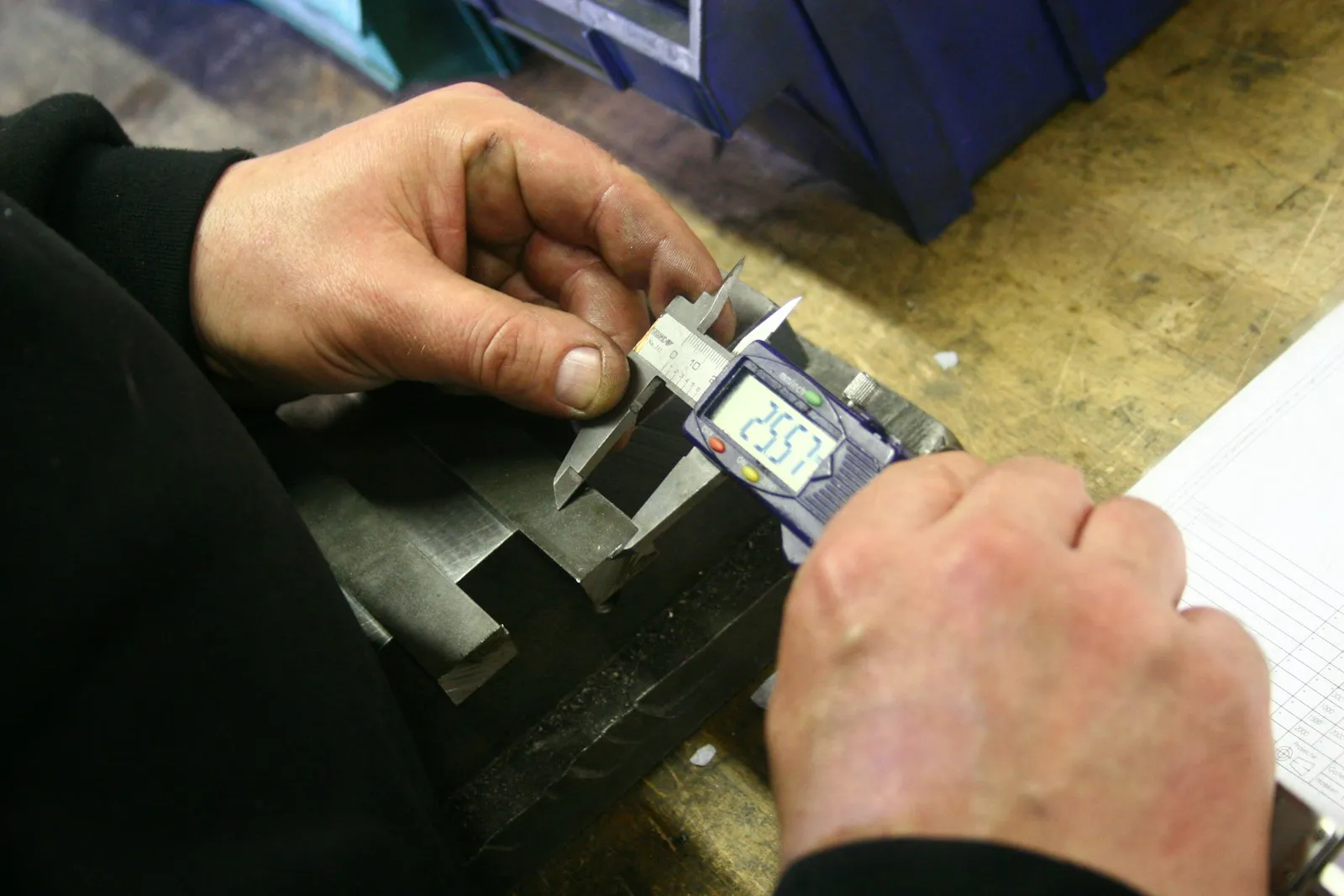 Manos midiendo una pieza metálica con un calibre digital en un banco de trabajo, con la pantalla marcando la medida