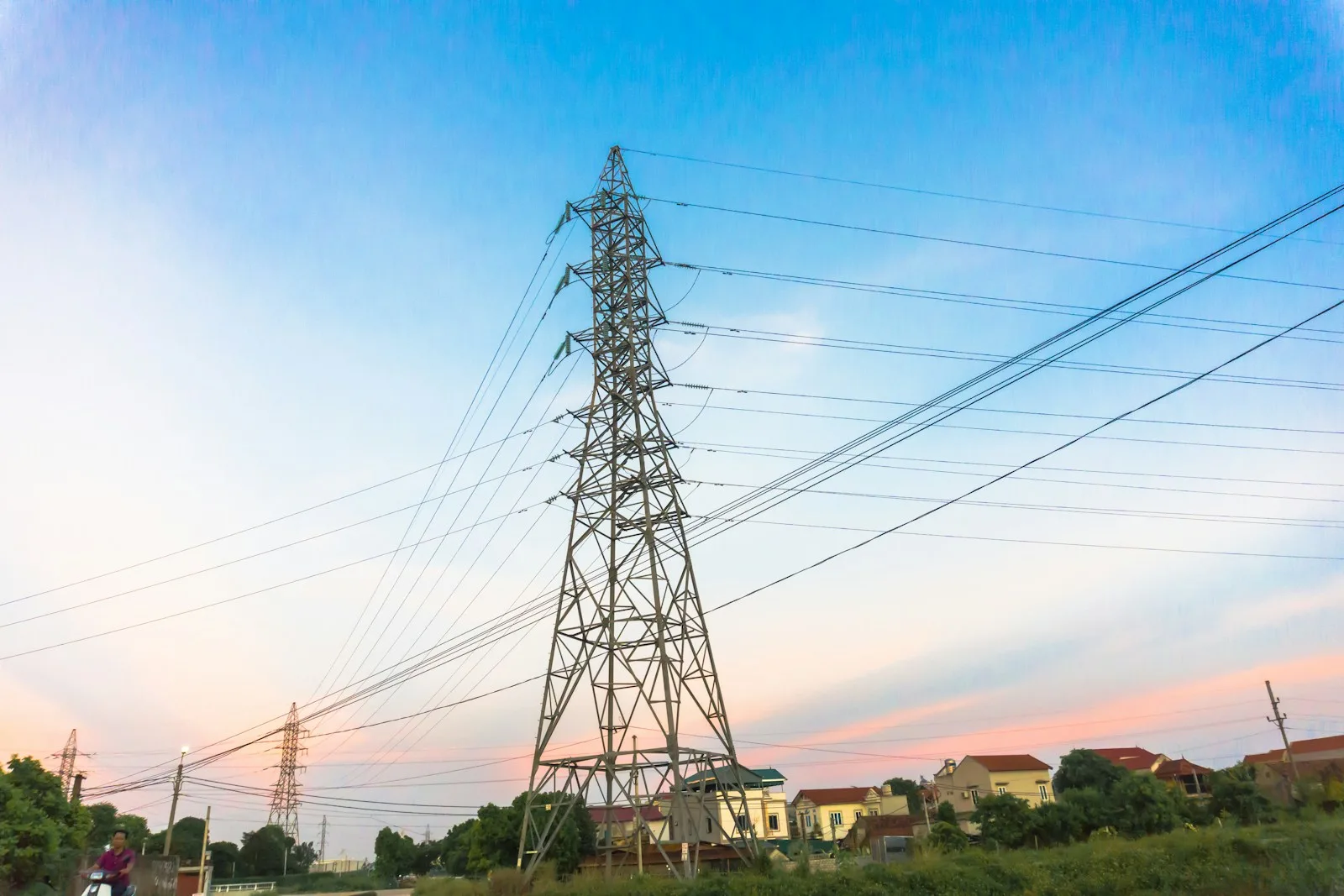 Torre de transmisión eléctrica con líneas que descienden hacia las casas de un pueblo al atardecer