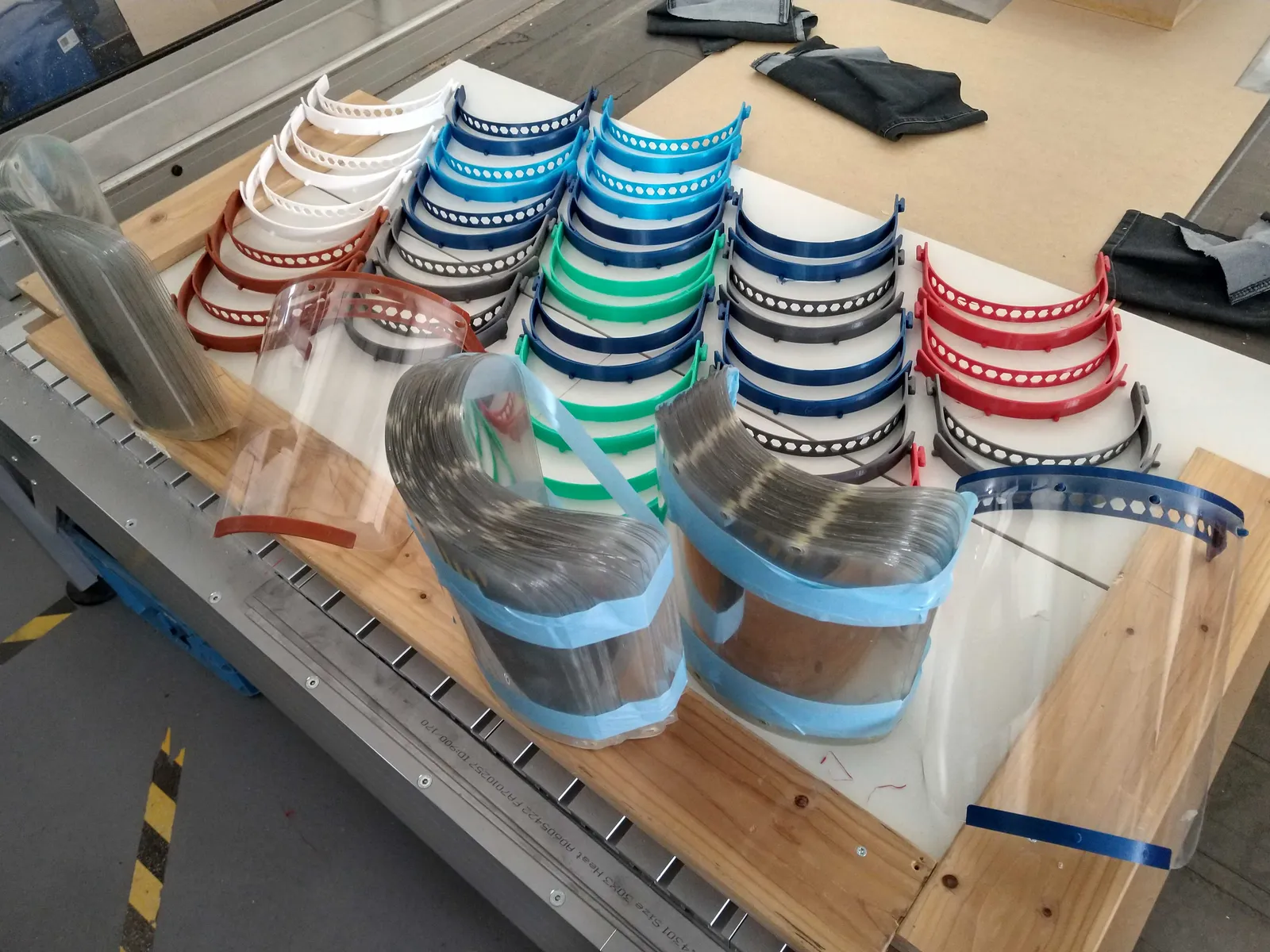 Decenas de diademas de visera impresas en 3D en varios colores, apiladas en un banco de trabajo junto a los plásticos transparentes de las pantallas