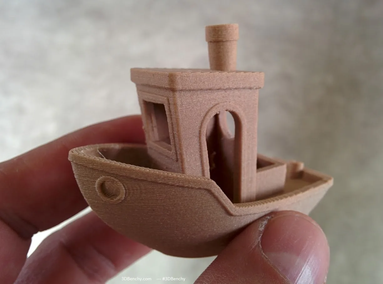 Barquito 3DBenchy impreso en filamento con relleno de madera, sostenido entre los dedos, de color tostado y mate