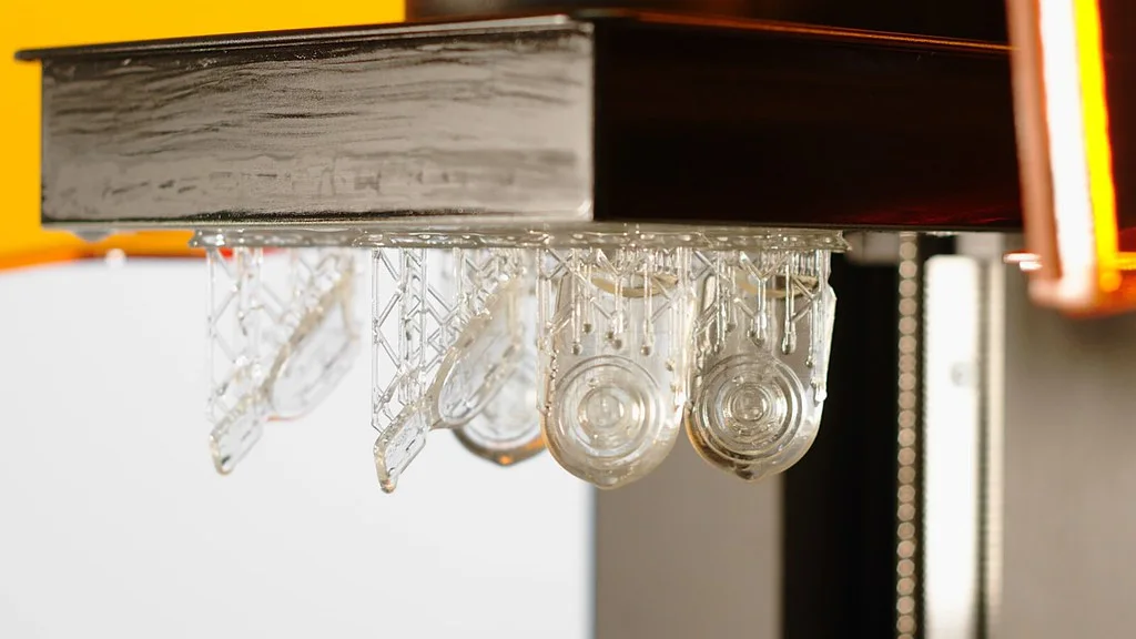 Piezas transparentes de resina colgando boca abajo de la plataforma de impresión, chorreando resina sin curar entre sus soportes