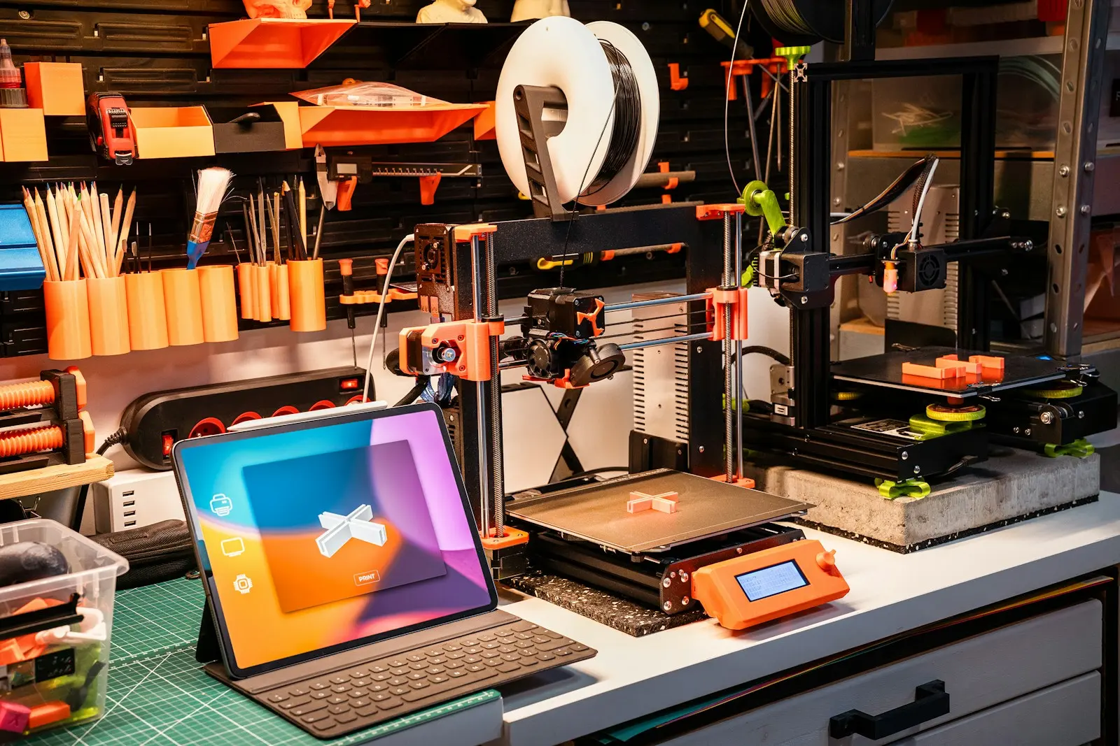 Dos impresoras 3D de estilos distintos trabajando en un banco de taller ordenado, con filamento y herramientas alrededor