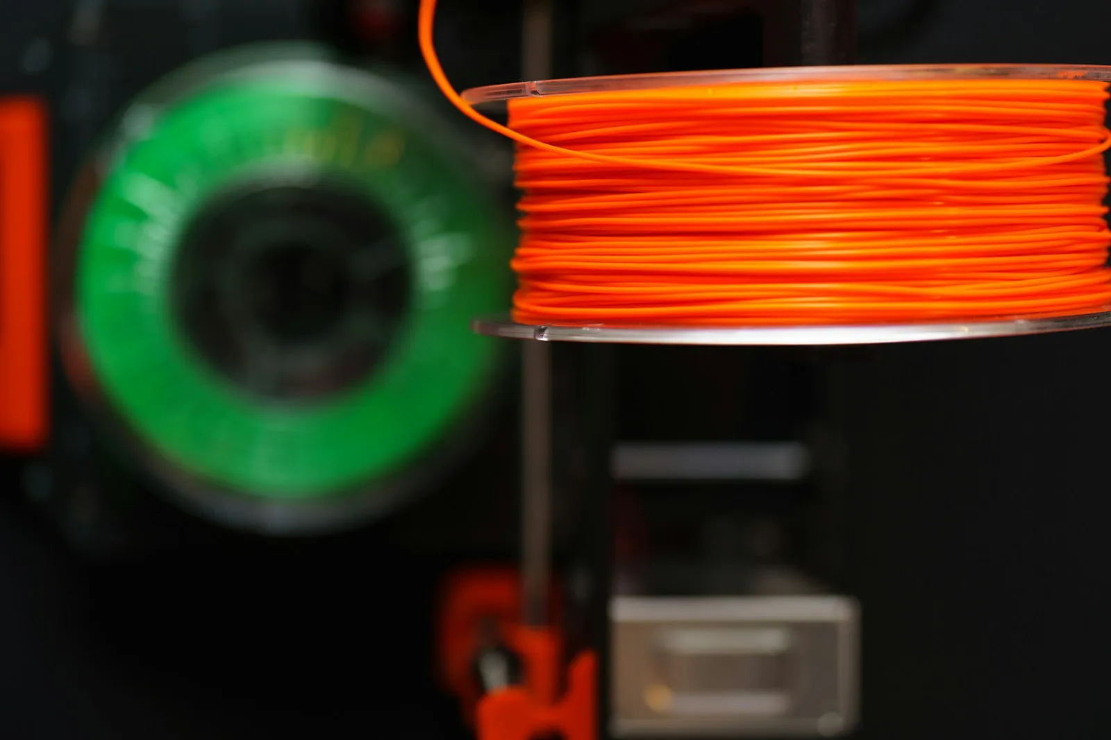 Bobina de filamento naranja en primer plano sobre una impresora, con otra bobina verde desenfocada al fondo