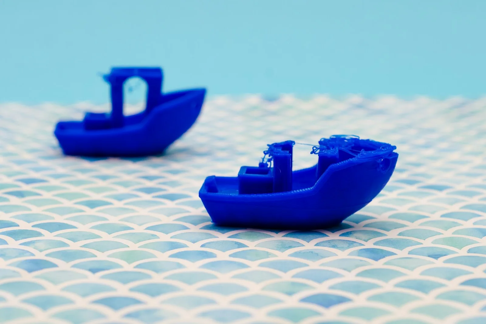 Dos barcos Benchy impresos en 3D en azul: uno limpio al fondo y uno con hilos y defectos de impresión en primer plano