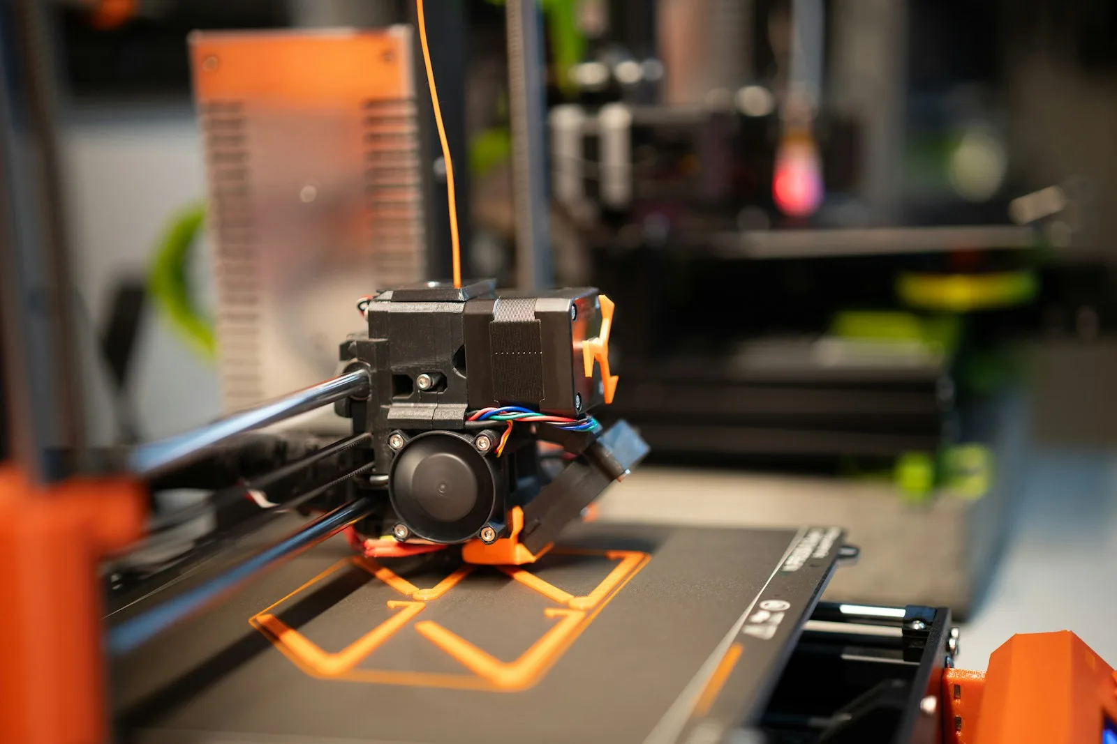 Cabezal de una impresora 3D depositando filamento naranja sobre la cama, con las primeras capas de la pieza ya trazadas