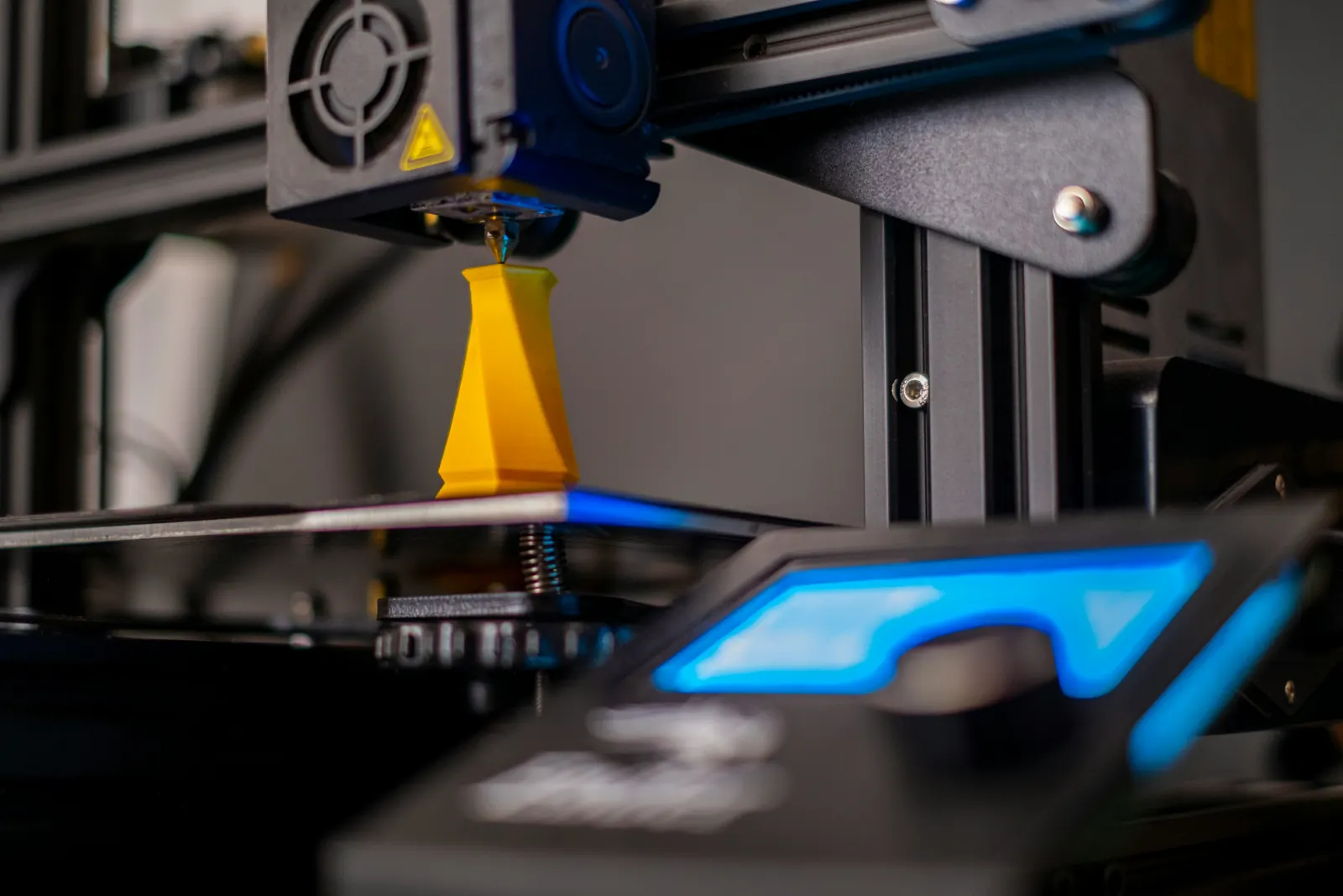 Cama de impresión negra con una pieza amarilla mientras la boquilla de una impresora 3D deposita material encima