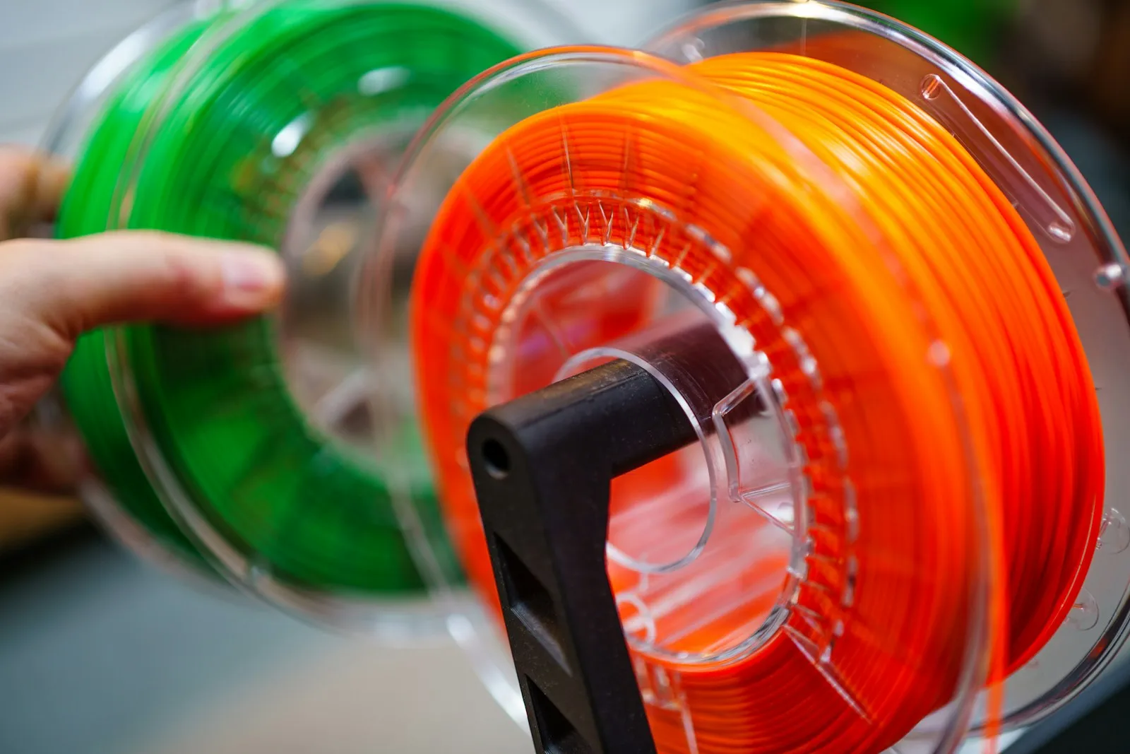 Una mano sostiene dos bobinas de filamento en carretes transparentes, una verde y otra naranja, con el bobinado a la vista