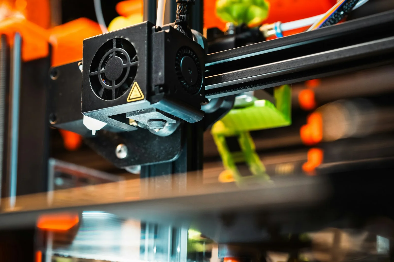 Cabezal extrusor de una impresora 3D en primer plano, con ventiladores y fondo naranja desenfocado